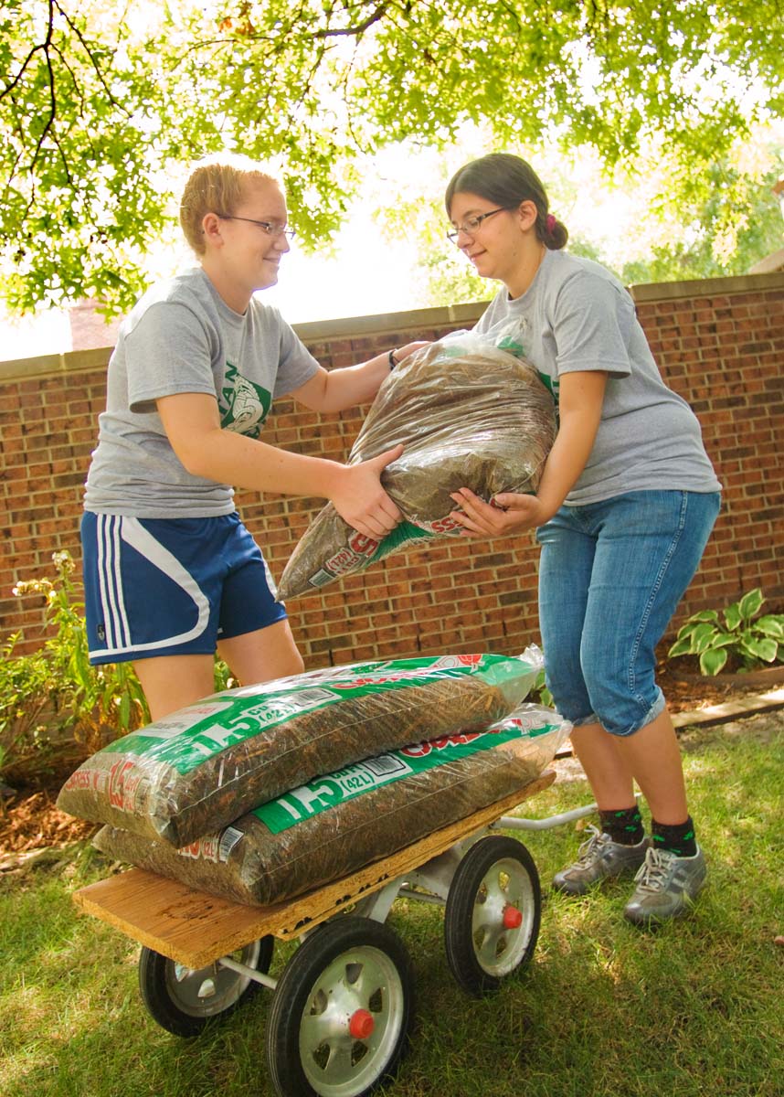 two students lift bag of mulch in yard