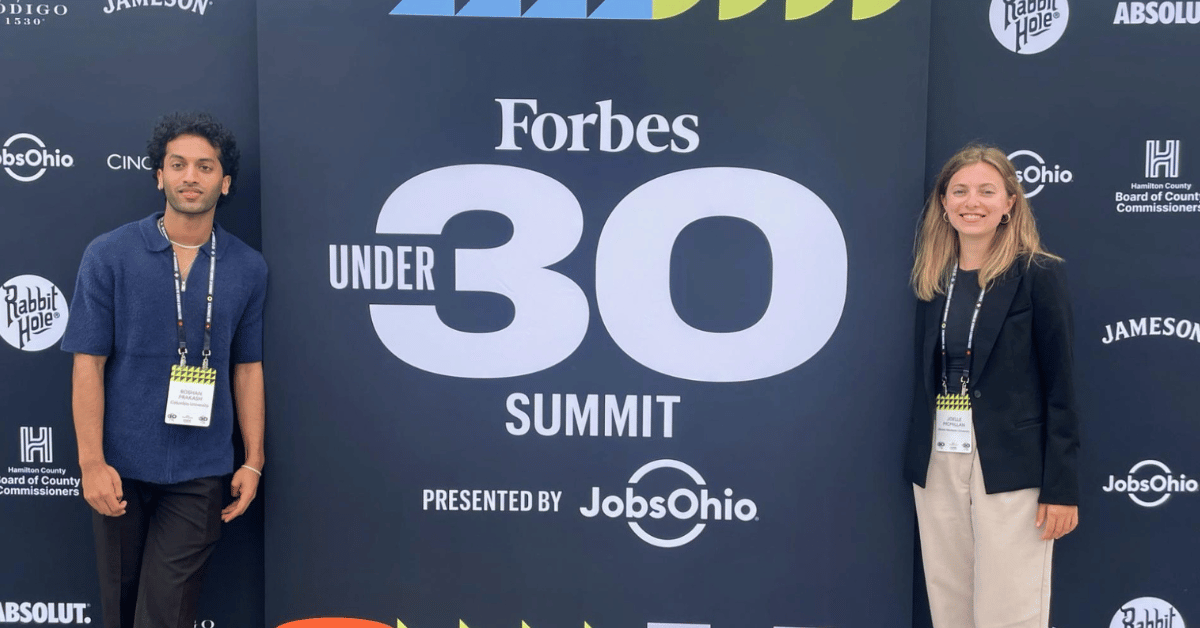 Roshan Prakash '24 and Joelle McMillan '25 stand by a banner at the Forbes Under 30 Summit in Cincinnati Ohio